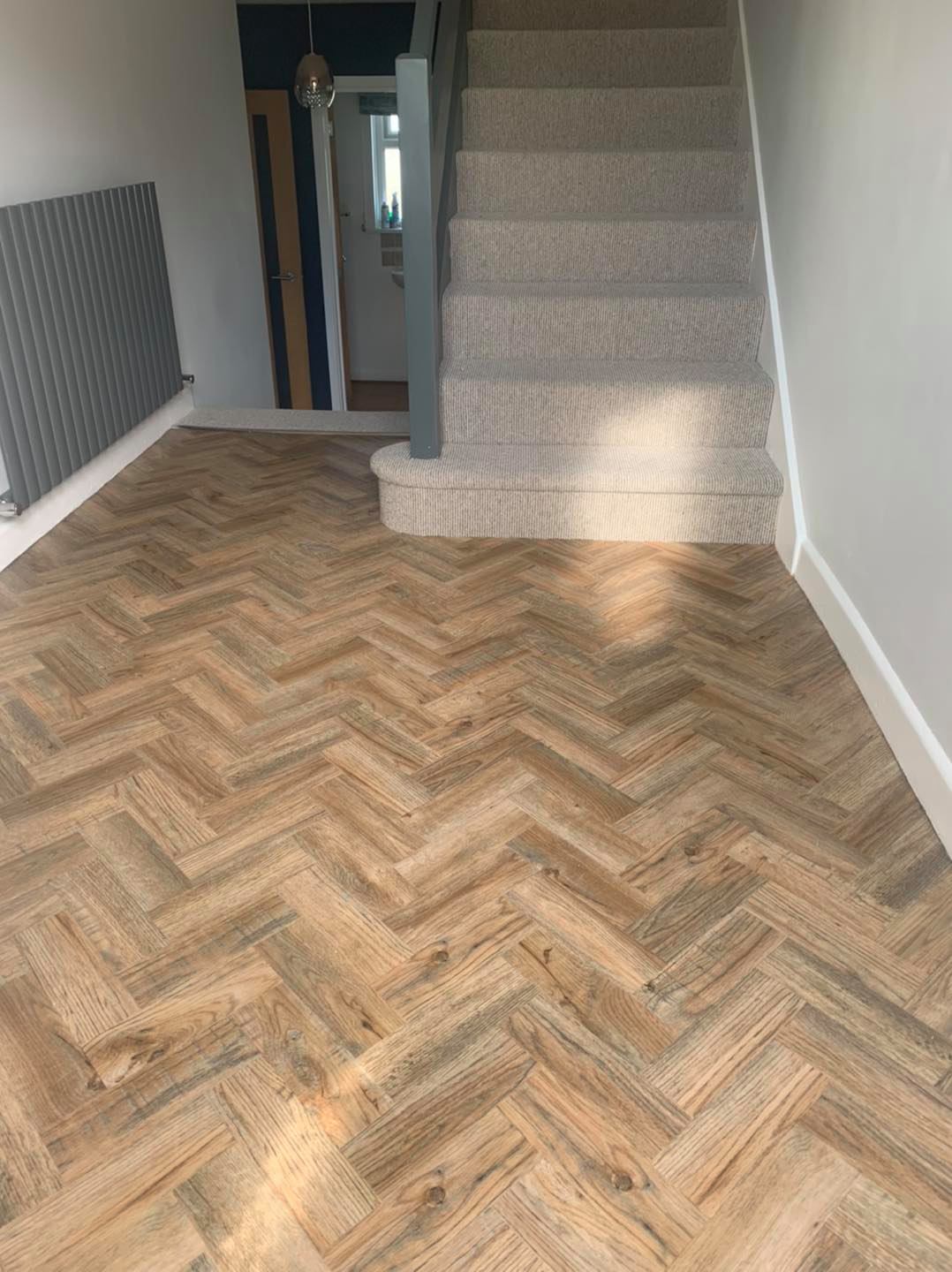 A hallway with a herringbone-patterned wooden floor leading to a carpeted staircase. A modern radiator is mounted on the left wall, and a door is visible at the end of the hallway.