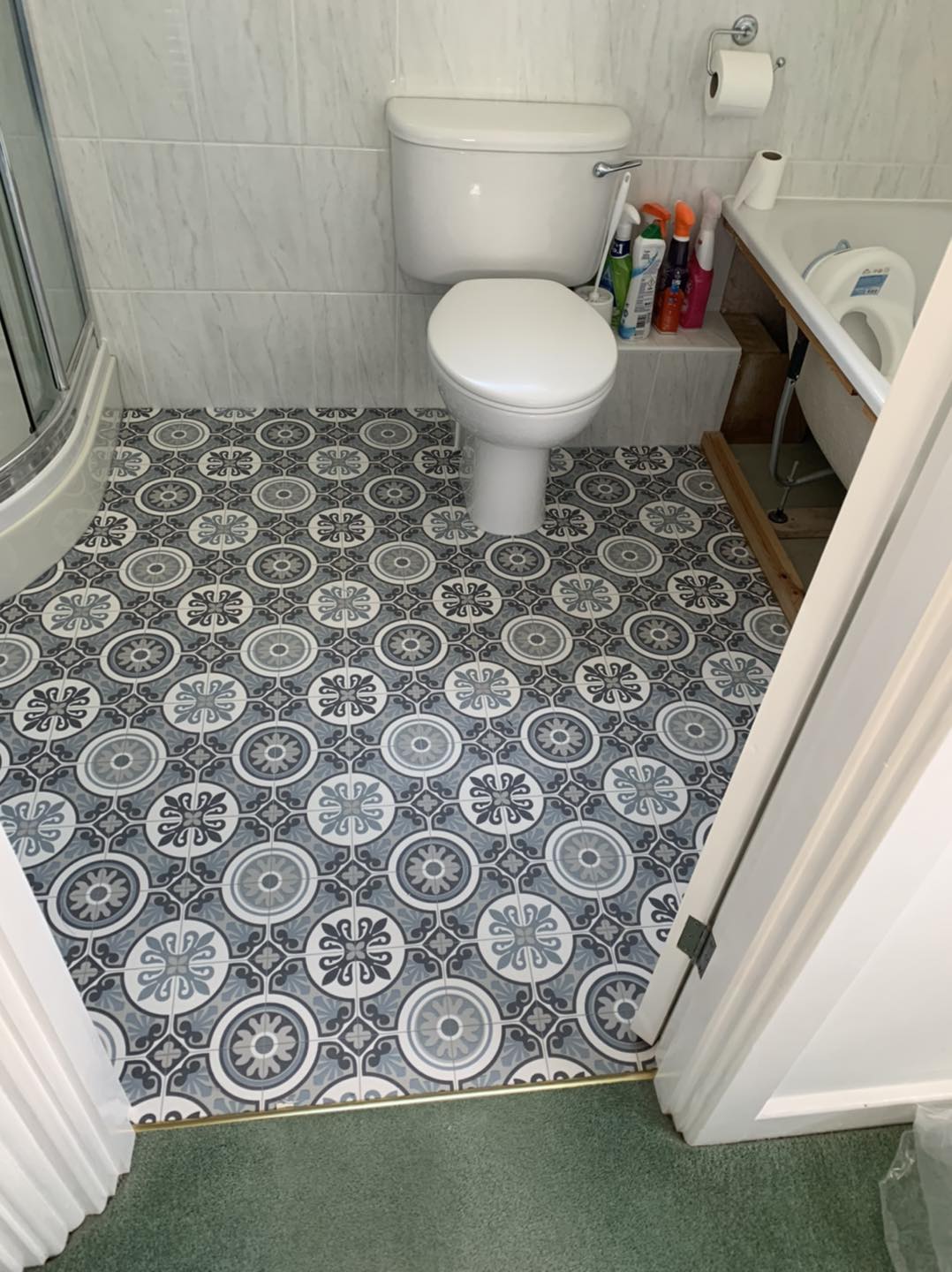 A small bathroom with patterned gray and white flooring, a white toilet, and a partially visible sink. Cleaning supplies are stored on a shelf under the sink, and a glass shower enclosure is situated in the corner.