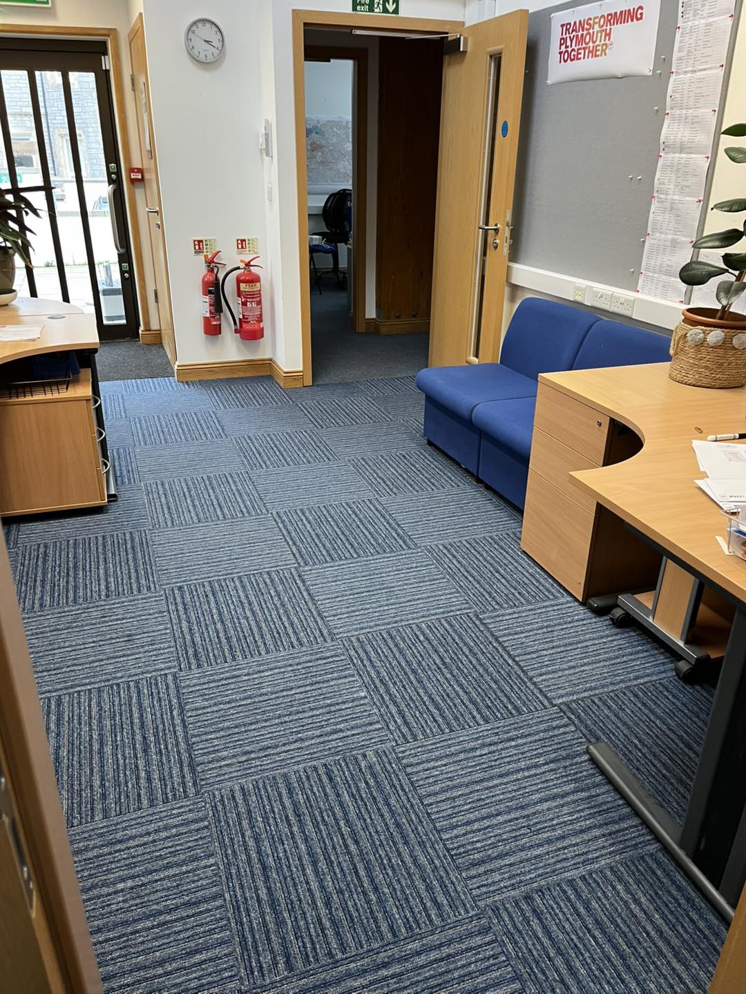 Office interior with blue patterned carpet, a blue couch, wooden desks, fire extinguishers, a wall clock, a potted plant, and an open doorway leading to another room. A poster is partially visible on the wall.
