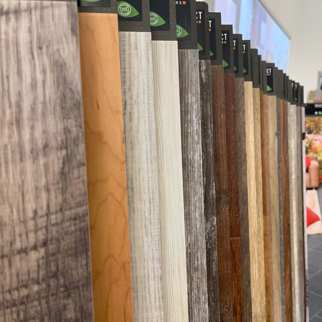 Close-up of various wood and laminate flooring samples in different colors and finishes, arranged vertically in a display rack inside a store.
