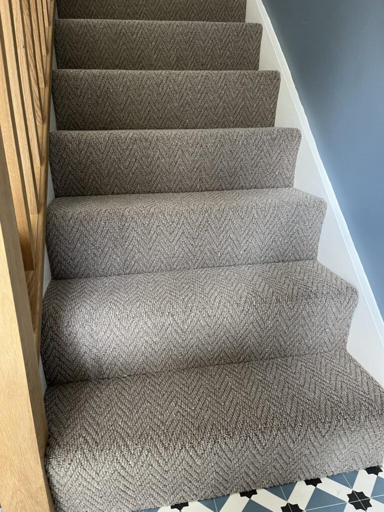 A staircase with a light brown patterned carpet runner, wooden railing on the left, white trim on the right, and geometric patterned floor tiles at the base.