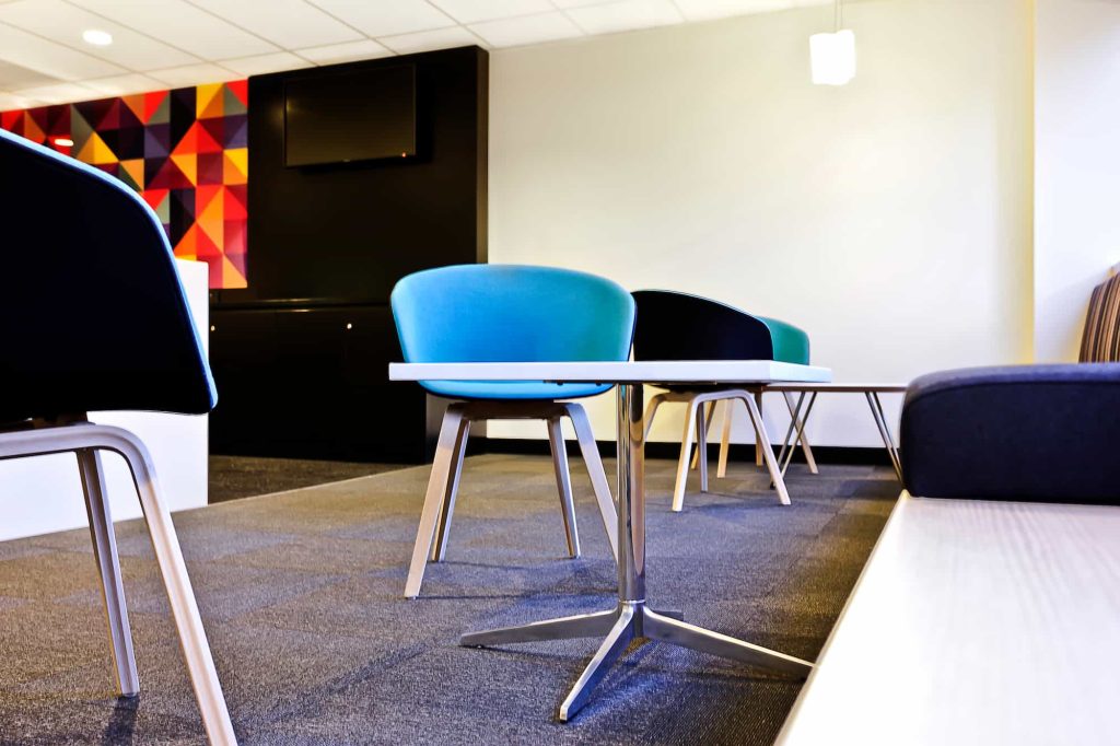 A modern waiting area with blue and black chairs around white tables, gray carpet flooring, a geometric colorful wall, and overhead lighting. The space looks clean and contemporary.