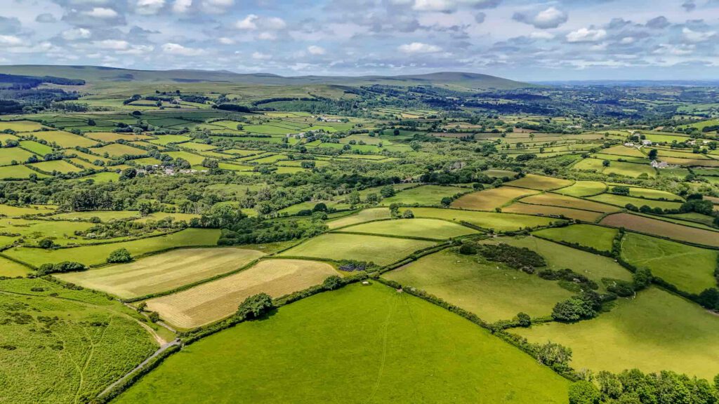 Aerial view of a patchwork of green and yellow fields, hedgerows, and scattered trees under a partly cloudy sky in a rural countryside landscape. Hills and distant valleys are visible on the horizon.