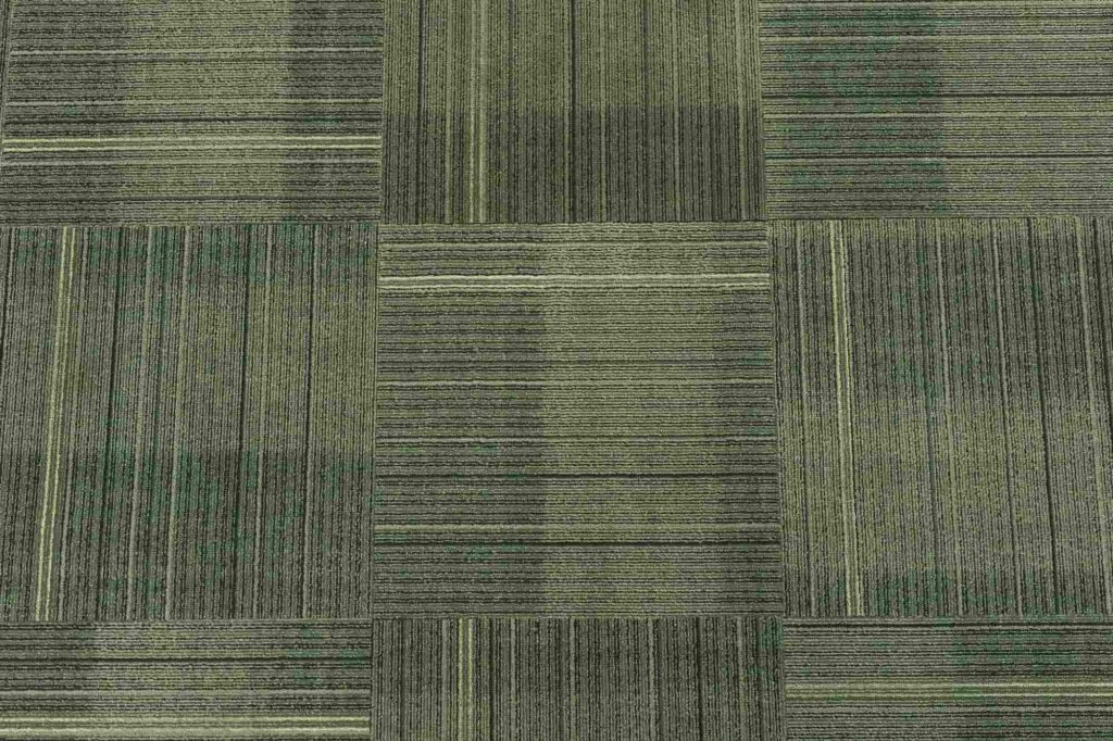 A pattern of square carpet tiles in varying shades of green and gray, each tile featuring subtle vertical and horizontal lines creating a textured, geometric, checkerboard effect.