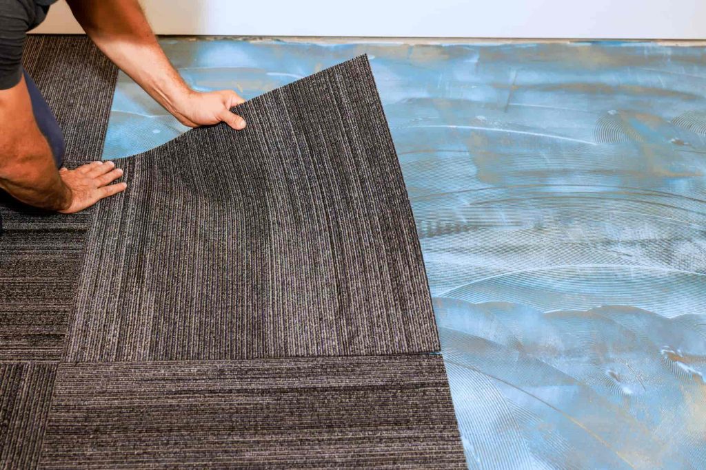A person’s hands are placing dark striped carpet tiles onto a floor covered in adhesive, installing new flooring in a room.