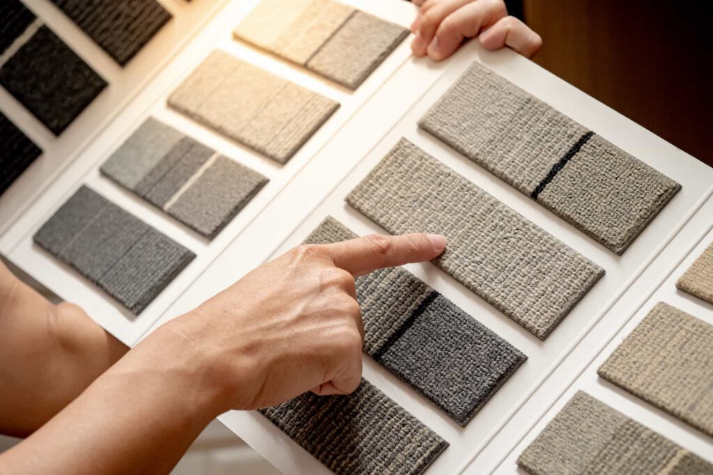 A person points to a textured gray carpet sample on a display board with several other carpet swatches in various shades of gray and beige. Another hand holds the board steady.