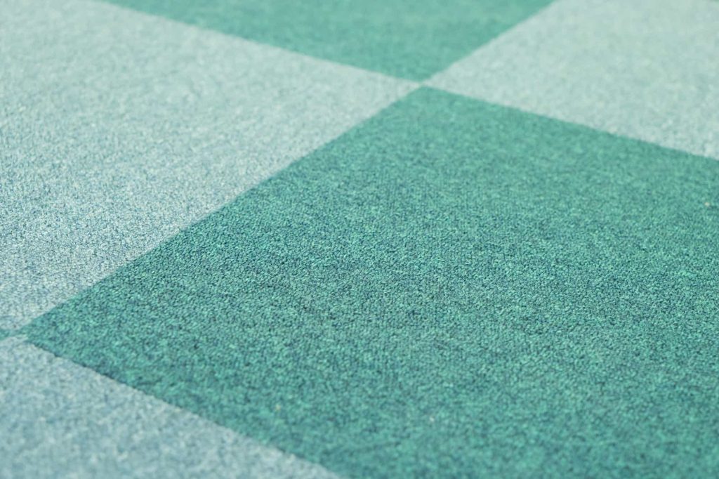 Close-up of a patterned carpet featuring large, alternating squares in different shades of blue and green, creating a checkered appearance. The texture of the carpet is visible.