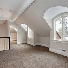 A bright attic room with sloped ceilings, arched windows, beige walls, and brown carpet flooring. Natural light enters through the windows, and a stair railing is visible on the left side.